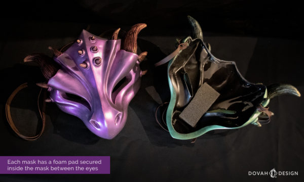 Two masquerade style draon masks sitting on a black table, one facing up, the other turned over to expose the black foam "nose padding" that comes affixed inside each mask.