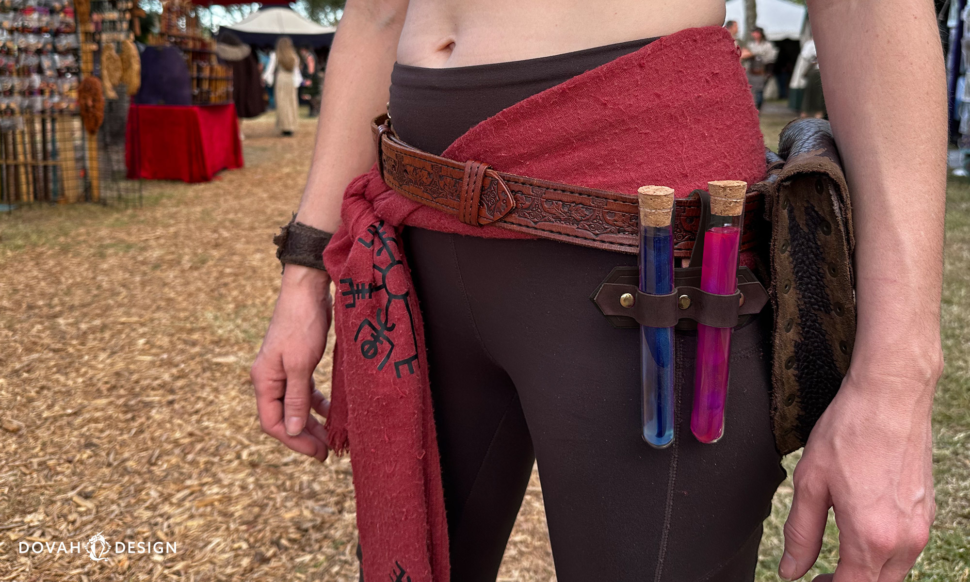 View of a "dark brown" double potion handmade leather holster affixed to a woman's belt, Renaissance Fair fair ground visible in the background. Potions seen in leather holster are blue and pink.