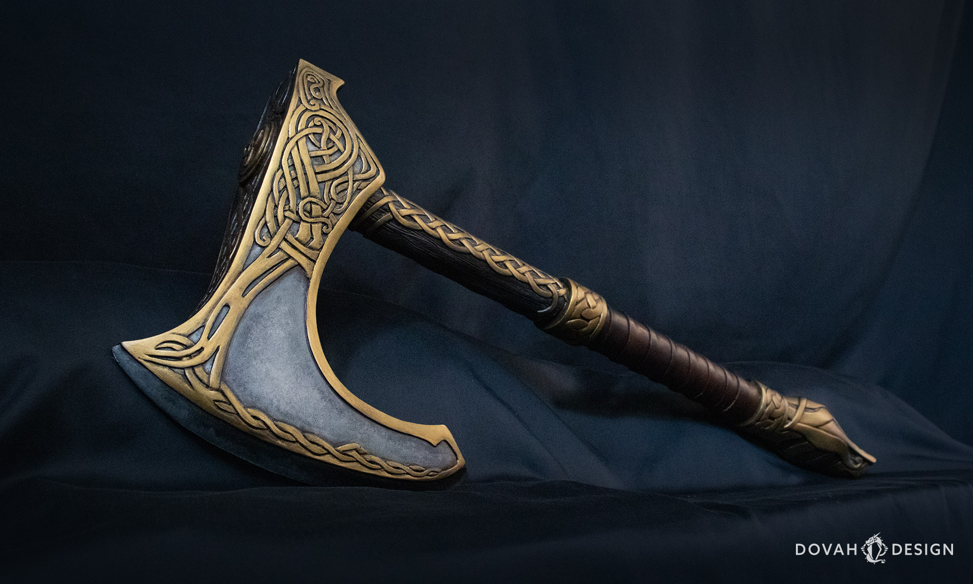 Angled view of Eivor's Raider Axe replica prop from Assassin's Creed: Valhalla, blade with golden knotwork detail in focus in the foreground, with knotwork hilt and handle seen farther in the distance in front of a black backdrop.