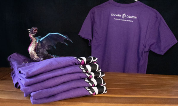 Stack of folded Dovah Design t-shirts sitting on a wooden table, a pruple toy dragon sitting atop the pile, with a hanging t-shirt behind showing the back design.