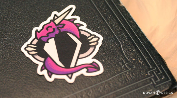 Dovah Design's logo, a purple dragon wrapped around an altered Skyrim shadowmark, printed as a dye-cut sticker. Pictured sitting on the corner of a Skyrim book.