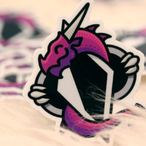 Dovah Design's logo, a purple dragon wrapped around an altered Skyrim shadowmark, printed as a dye-cut sticker. Pictured sitting upright on a sheepskin, with more stickers scattered in the background.