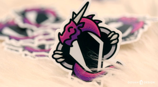 Dovah Design's logo, a purple dragon wrapped around an altered Skyrim shadowmark, printed as a dye-cut sticker. Pictured sitting upright on a sheepskin, with more stickers scattered in the background.