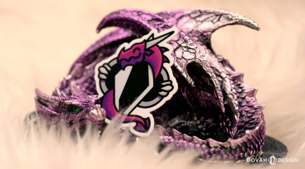 Dovah Design's logo, a purple dragon wrapped around an altered Skyrim shadowmark, printed as a dye-cut sticker. Pictured leaning up against a purple dragon statuette.