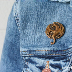 Elsweyr dragon logo lapel pin in gold, pinned on a jean jacket for scale, close up detail shot of the cast resin pin.