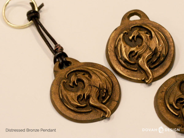 Distressed bronze pendant of the Elder Scrolls: Online Elsweyr logo, shown tied as a keychain, keyring, lying flat. Logo depecits a drake (dragon) symbol, purposely aged to look like old treasure.