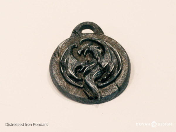 Distressed iron resin cast pendant of the Elder Scrolls: Online Elsweyr logo, shown lying flat. Logo depecits a drake (dragon) symbol, purposely aged to look like old treasure.