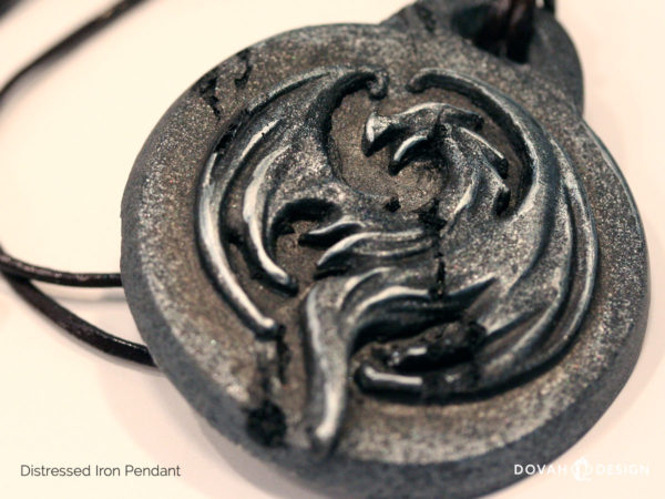 Close up detail of the distressed iron pendant of the Elder Scrolls Online Elsweyr logo. Pendant depicts a drake, curled in to a circle. Pendant has been distressed to look like old treasure.