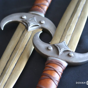 Close up detail of two calcinium (gold) khajiit dagger props, design based on the Elder Scrolls Online. Both crossguards are curved like a crescent moon, with four-pointed star on top.