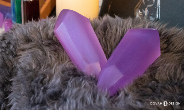 Close up detail of two lesser soul gems of slightly varying pink-purple color, resting atop gray fur with a Skyrim themed set in the background.