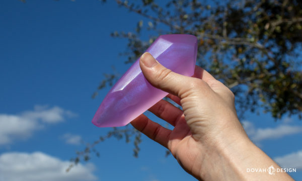 A single lesser soul gem being held in a hand outdoors in front of a blue sky, metallic powder seen glittering within the resin gem in the sunlight.