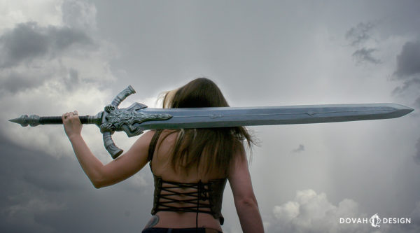 Sam holding her prop Greatsword of Artorias horizontally over her left shoulder, and across her back to the right shoulder. A cloudy gray sky in the background,