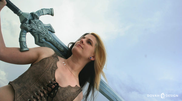 Sam looks off to the left, holding the greatsword of Artorias prop over her right shoulder, camera angled upward with a bright blue sky in the background.