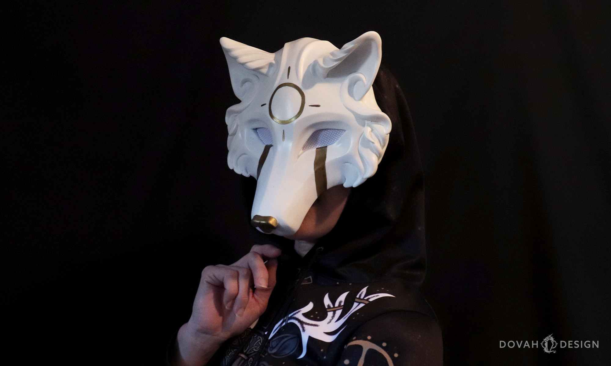 White "Skoll" wolf mask worn by a woman in a black hoodie, posed facing to the left in front of a black background.