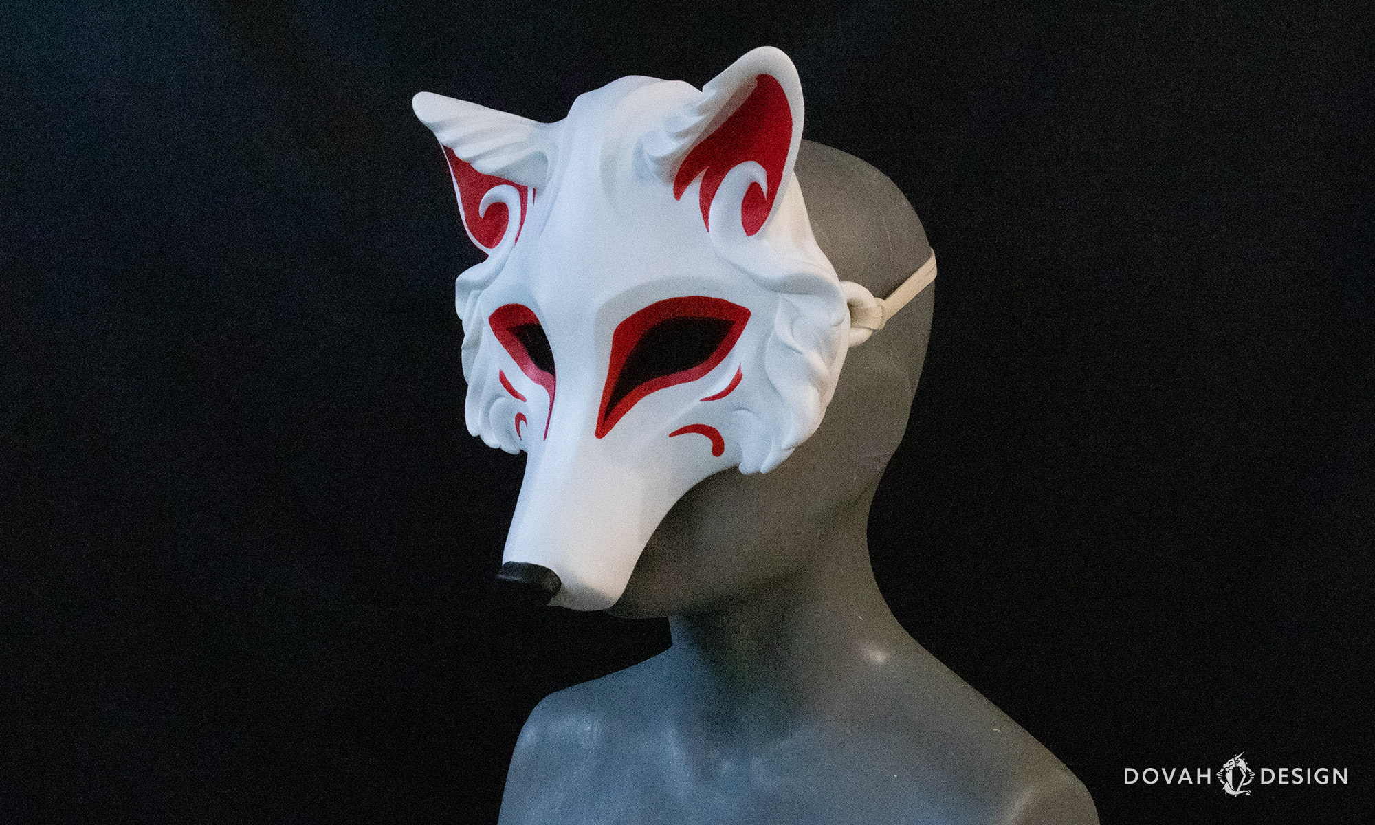 Side angle view of a white wolf mask painted in the Japanese Kistune-style with red eye and facial designs and a black nose, posed on a mannequin.