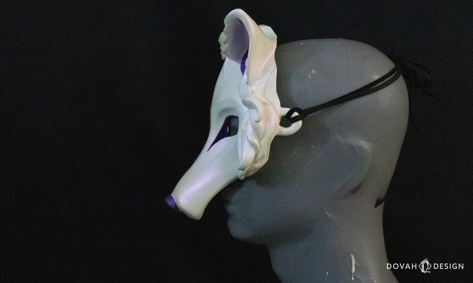 Side left view of a white wolf mask with purple accents, tied onto on a mannequin bust with black leather lace in front of a black background.